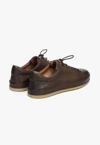 Hnědé kožené tenisky s nízkým profilem, kulatou špičkou a minimálním šněrováním. Mají béžovou podšívku a béžovou gumovou podrážku. Žádné viditelné vzory.