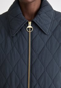 Κουβέρτα μπουφάν σε σκούρο ναυτικό χρώμα με διαμαντένιο σχέδιο, διαθέτοντας χρυσό φερμουάρ και μεγάλο γιακά, κατασκευασμένο από υφή υφάσματος.