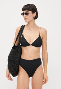 Black ribbed bikini with a triangular top and high-waisted bottom. Model holds a black woven tote bag. Accessories include sunglasses.