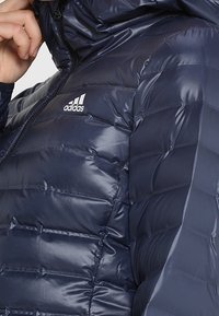 Veste matelassée bleu marine avec une texture brillante et matelassée. Dispose d'une fermeture éclair à l'avant, d'une capuche et du logo Adidas blanc sur la poitrine.