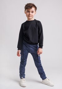 Młody chłopiec stoi z nogami rozstawionymi, ubrany w czarną bluzę, niebieskie spodenki w wzory z kieszeniami i białe sportowe buty na jednolitym tle.