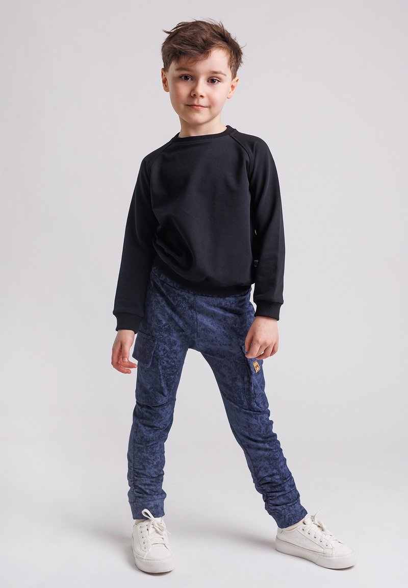 Młody chłopiec stoi z nogami rozstawionymi, ubrany w czarną bluzę, niebieskie spodenki w wzory z kieszeniami i białe sportowe buty na jednolitym tle.