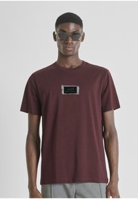 Camiseta de algodón color marr ón con cuello redondo, que presenta un parche de logo negro texturizado en el pecho. El modelo lleva pantalones grises y gafas de sol oscuras.