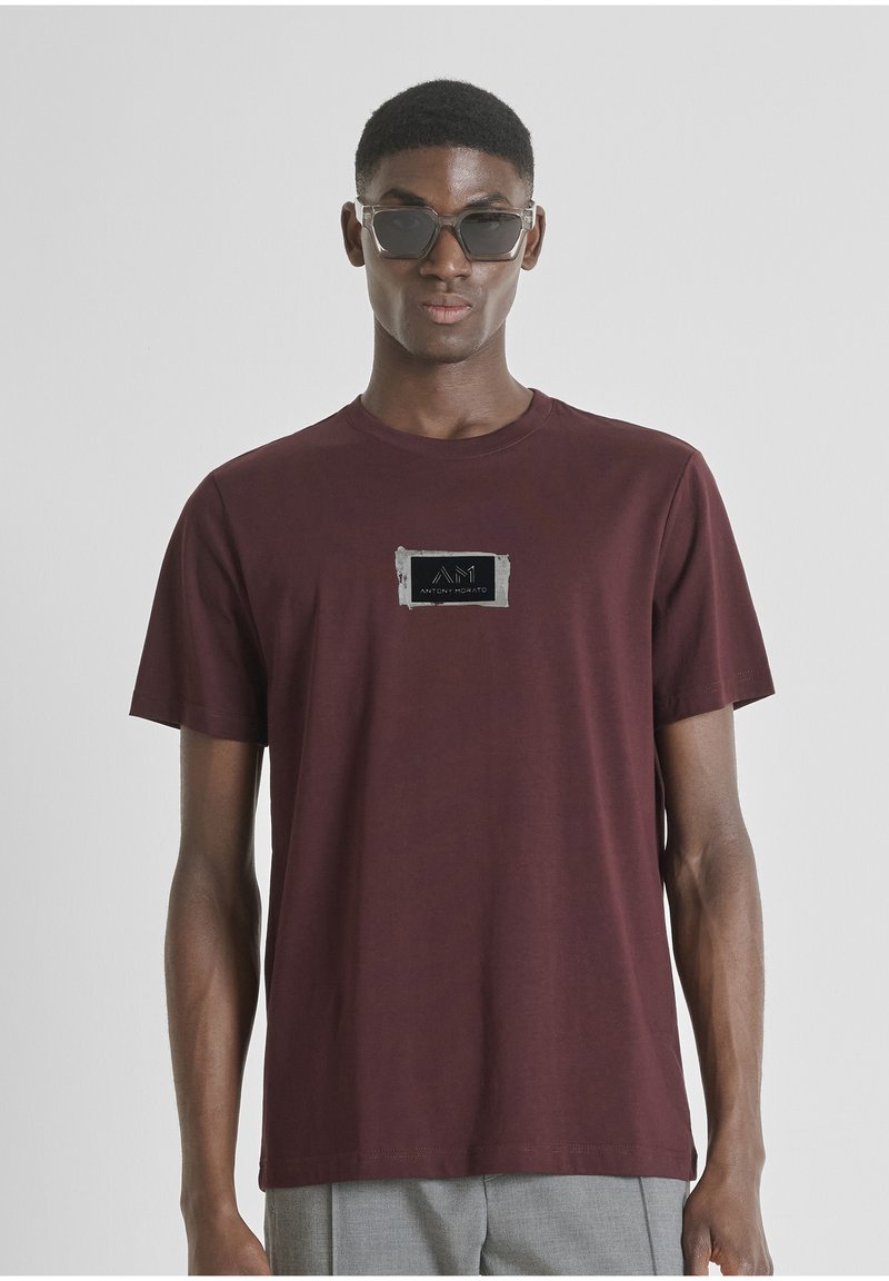 Camiseta de algodón color marrón con cuello redondo, que presenta un parche de logo negro texturizado en el pecho. El modelo lleva pantalones grises y gafas de sol oscuras.