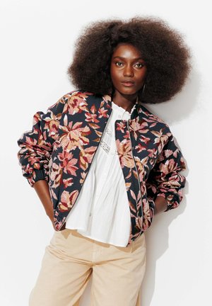 Femme aux cheveux afro naturels portant une veste bomber à motif floral sur un chemisier blanc et un pantalon beige, debout devant un fond blanc.