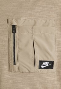Harmaa kangaspaita, jossa on teksturoitu pinta, suuri läpällinen tasku mustalla vetoketjulla sekä musta Nike-logo.