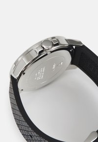 Schwarzes Silikon-Uhrenarmband mit grauem Logomuster, silbernes Edelstahlgehäuse, kreisförmige Form und auffällige Seitenkrone.