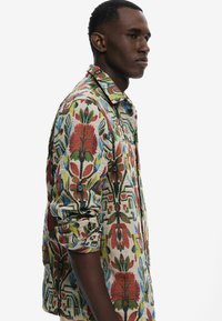 Bunt gemusterte Button-Up-Bluse mit floralen Designs in Rot, Grün und Blau auf einem beigen Hintergrund. Klassischer Kragen und hochgekrempelte Ärmel.
