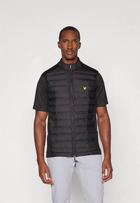 Mann trägt eine schwarze kurzärmlige Steppweste mit gelbem Adlerlogo, schwarzes T-Shirt und hellgraue Hose, steht vor einem schlichten Hintergrund.