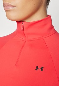 Rød idrettsjakke med høy krage, med svart Under Armour-logo på venstre side. Laget av glatt, elastisk stoff.
