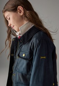 Mørkeblå voksbehandlet lærredsjakke med en krave i kordfløjl, gyldne metalaccenter og en frontlomme med et synligt logo-patch.