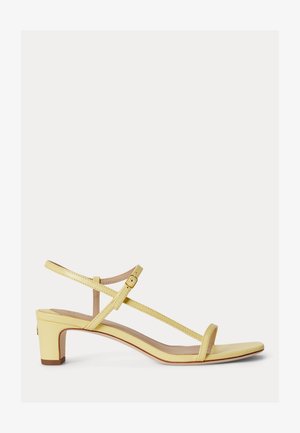 Rumene sandale z nizko peto, tankimi trakovi, zaponko ob gležnju in odprtim prstom na belem ozadju.