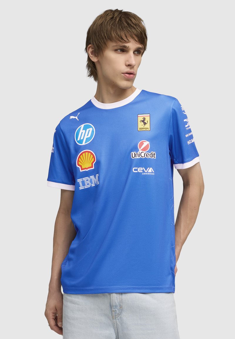 Ung mand iført en blå Ferrari teamtrøje med sponsorskaber (HP, Shell, IBM, UniCredit, CEVA) og hvide ærmet trims, der ser til venstre.