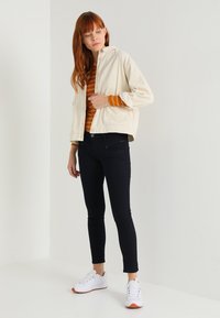 Chaqueta de sarga color crema con diseño corto, camiseta rayada en naranja y blanco, jeans ajustados negros y zapatillas blancas con detalles en naranja.