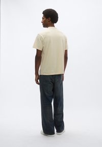 Μπεζ κοντομάνικη μπλούζα, σε χαλαρή γραμμή, συνδυασμένη με σκούρο μπλε τζιν παντελόνι με φαρδιά πόδια και λευκά αθλητικά παπούτσια, σε προοπτική από πίσω πάνω σε ουδέτερο φόντο.