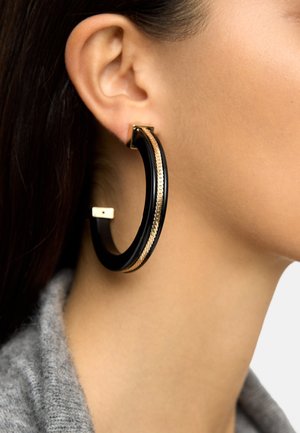 Zbliżenie na ucho kobiety noszącej duży czarny kolczyk-koło z detalem z złotego łańcuszka, na tle szarego swetra i jasnego tła.