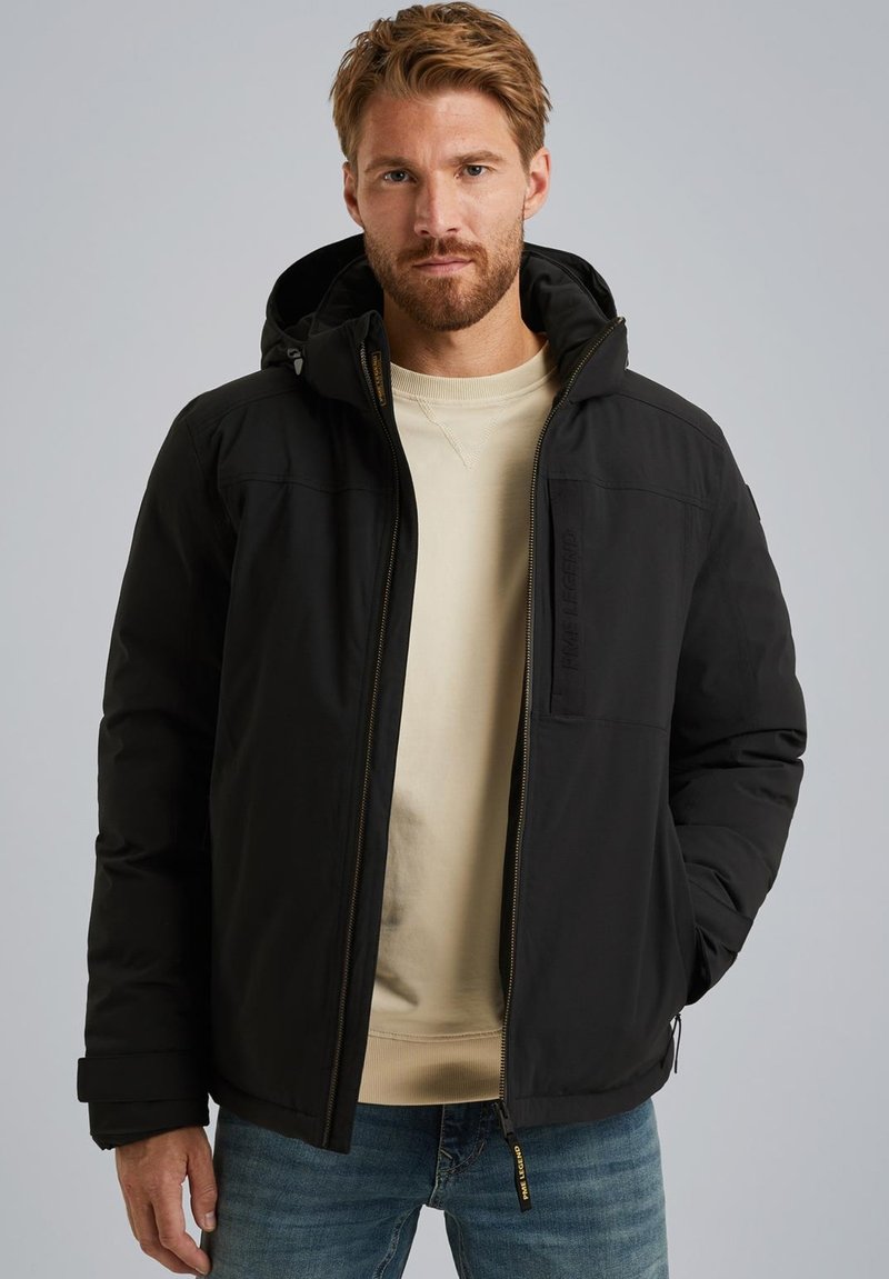 Giacca nera isolata con cappuccio, dotata di chiusura con zip, tasche laterali e tessuto texture. Indossata sopra un felpa beige chiaro e jeans blu.