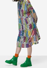 Kleurrijke knielange jurk met een huisprint, gecombineerd met felgroene enkellaarzen en oranje geribbelde sokken. Gladde, lichte stof.
