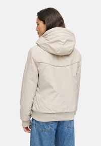 Beige pufferjack met kap, voorzien van ribgebreide manchetten en zoom, een soepele textuur en subtiele stiksels op de achterkant.