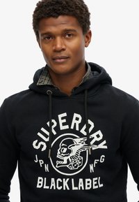 Sudadera negra con un gráfico texturizado en blanco de un cráneo con alas y dados. Cuerdas de ajuste con herrajes plateados y una camisa de cuadros en capas por debajo.