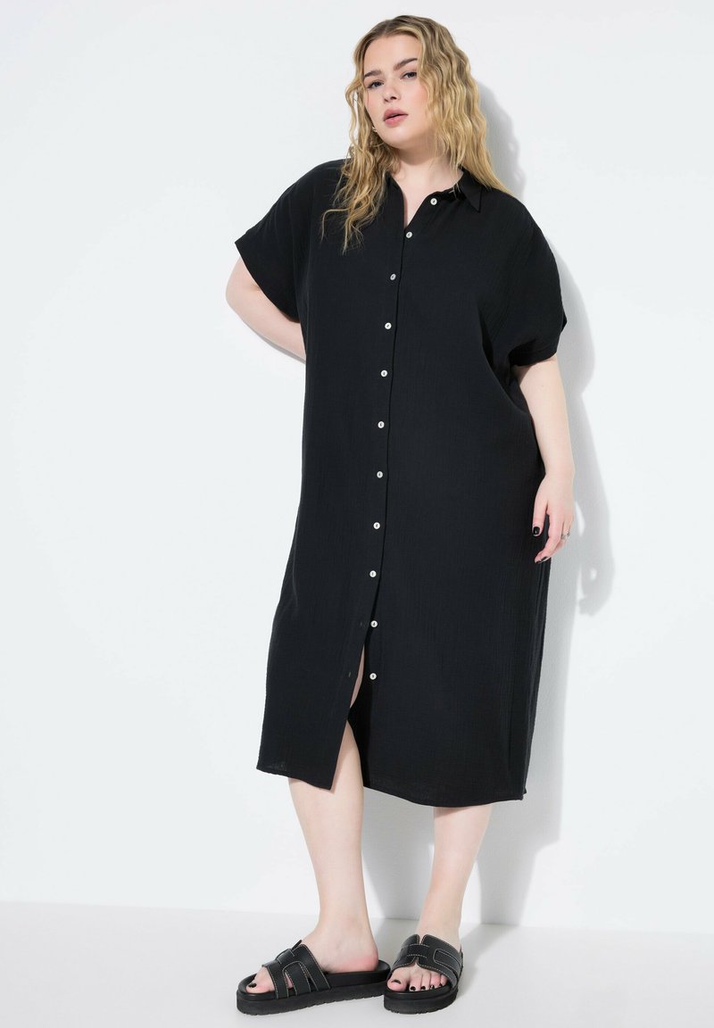 Vestido negro con botones, de manga corta y ajuste relajado, en tela texturizada, con una abertura frontal, combinado con sandalias negras tipo slide.