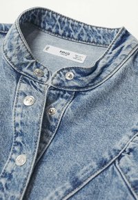 Veste en jean bleu clair avec un col mandarin et trois boutons-pression argentés visibles le long de la patte de boutonnage avant.