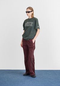 Prevelika zelena tie-dye Nike majica s bijelim logom, u kombinaciji s burgundskim hlačama s bijelim obrubom i crnim cipelama.