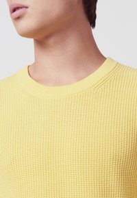 Gelber gestrickter Pullover mit Rundhalsausschnitt, der ein strukturiertes Muster und gerippte Bündchen aufweist. Nahaufnahme des Kragens und des Oberbrustbereichs.