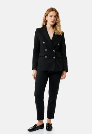 Frau mit welligem blondem Haar, die einen schwarzen doppelreihigen Blazer, schwarze Hose und schwarze Loafers trägt, steht vor einem einfarbigen Hintergrund.