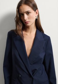 Giovane donna con lunghi capelli castani, che indossa un blazer blu scuro con scollo a V profondo e orecchini a cerchio d'argento, in piedi contro una parete chiara e semplice.