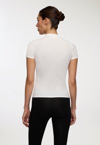 Biała bluzka z krótkim rękawem o teksturowanym, prążkowanym wykończeniu. Posiada wysoki dekolt i dopasowany krój, zestawiona z czarnymi legginsami.