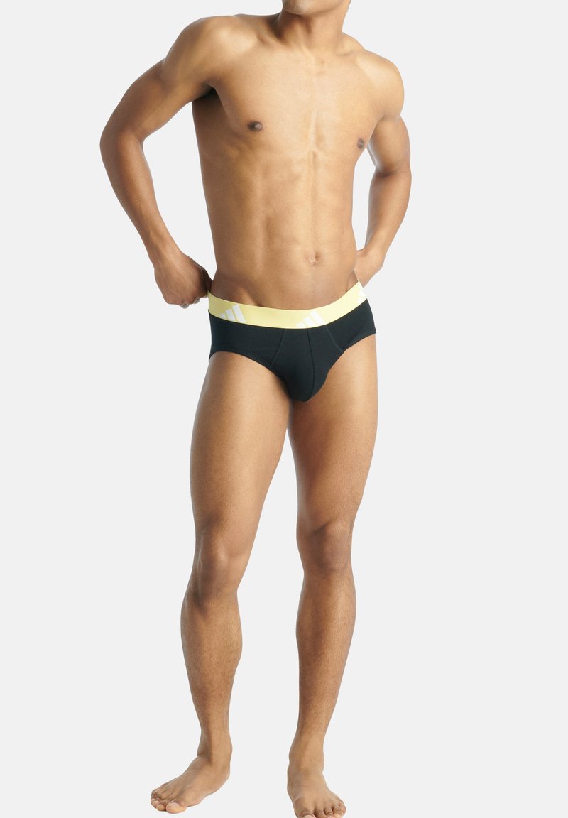 Schwarze Badehose mit gelbem Bund, der diagonale weiße Streifen aufweist, aerodynamisches Design und glatte Textur.