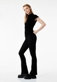 Top negro ajustado con mangas cortas combinado con pantalones acampanados negros. La modelo lleva unos zapatos de plataforma gruesos y negros. Tela suave y elástica.