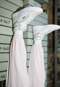 Piernas en pantalones de chándal rosa elevadas sobre cajas de zapatos apiladas, llevando zapatillas Puma blancas y en tonos pastel con logotipos y cordones visibles.