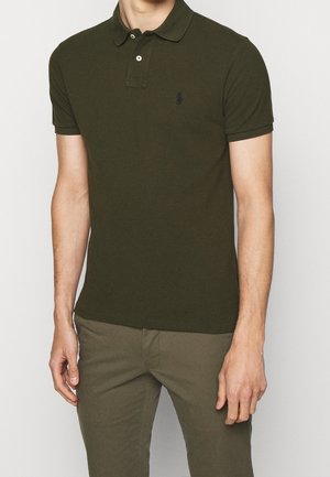 Homme portant un polo vert foncé à manches courtes avec deux boutons et un petit logo brodé sur la poitrine, associé à un pantalon olive assorti.