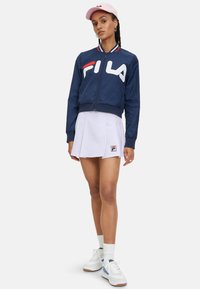 Marineblauer Bomberjacke mit großem weißen Fila-Logo, kontrastierenden roten und weißen Kragenstreifen, kombiniert mit einem gefältelten weißen Rock und weißen Sneakers.