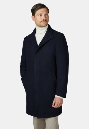 Mann trägt einen dunkelblauen Mantel über einem weißen Rollkragenpullover und beigen Hosen, schaut zur Seite mit teilweise in den Manteltaschen steckenden Händen.