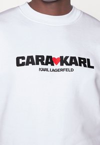 Sudadera blanca con el texto negro en negrita "CARA❤️KARL" y "KARL LAGERFELD" en letras más pequeñas. Tejido suave con diseño de cuello redondo.