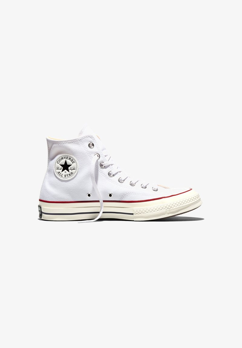 Bílá vysoká teniska Converse Chuck Taylor All Star s černým logem hvězdy a červeným pruhem na podrážce, zobrazena z boku.