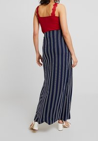 Rode tanktop met kantaccenten, gecombineerd met een marineblauwe maxrok met witte verticale strepen. De outfit wordt afgemaakt met witte blokhak sandalen.