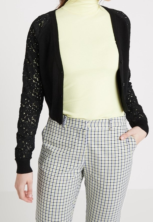 Svart kofta med spetsärmar, knapplock, bärs över en ljusgul ribbad turtleneck, ihopparad med rutiga byxor.