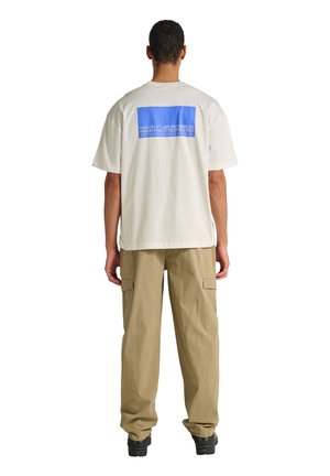 Mężczyzna noszący białą, oversize'ową koszulkę z niebieskim prostokątnym napisem na plecach oraz beżowe spodnie typu cargo z czarnymi butami, stojący tyłem.