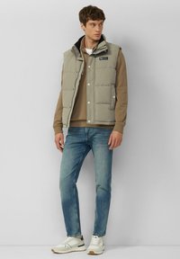 Lichtgrijze gewatteerde bodywarmer met zijzakken, drukknopen en een hoge kraag, gedragen over een beige sweatshirt, gecombineerd met blauwe jeans en witte sneakers.