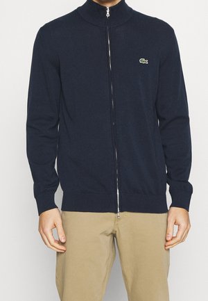 Homme portant un cardigan zippé bleu marine avec un logo de crocodile vert brodé sur la poitrine et un pantalon beige, visible du cou jusqu'aux hanches.