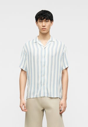 Jeune homme portant une chemise blanche à manches courtes avec des rayures verticales bleues et un pantalon beige, debout devant un fond clair uni.