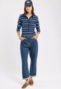 Gestreiftes Langarmhemd in Marineblau, Türkis und Weiß. Blaue Jeans mit umgeschlagenen Säumen. Braune Bootsschuhe. Blaue Kappe mit Logo.