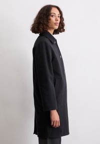 Eine Frau mit lockigen Haaren trägt einen schwarzen, knopfartigen Mantel mit Taschen und steht seitlich vor einem schlichten, hellgrauen Hintergrund.