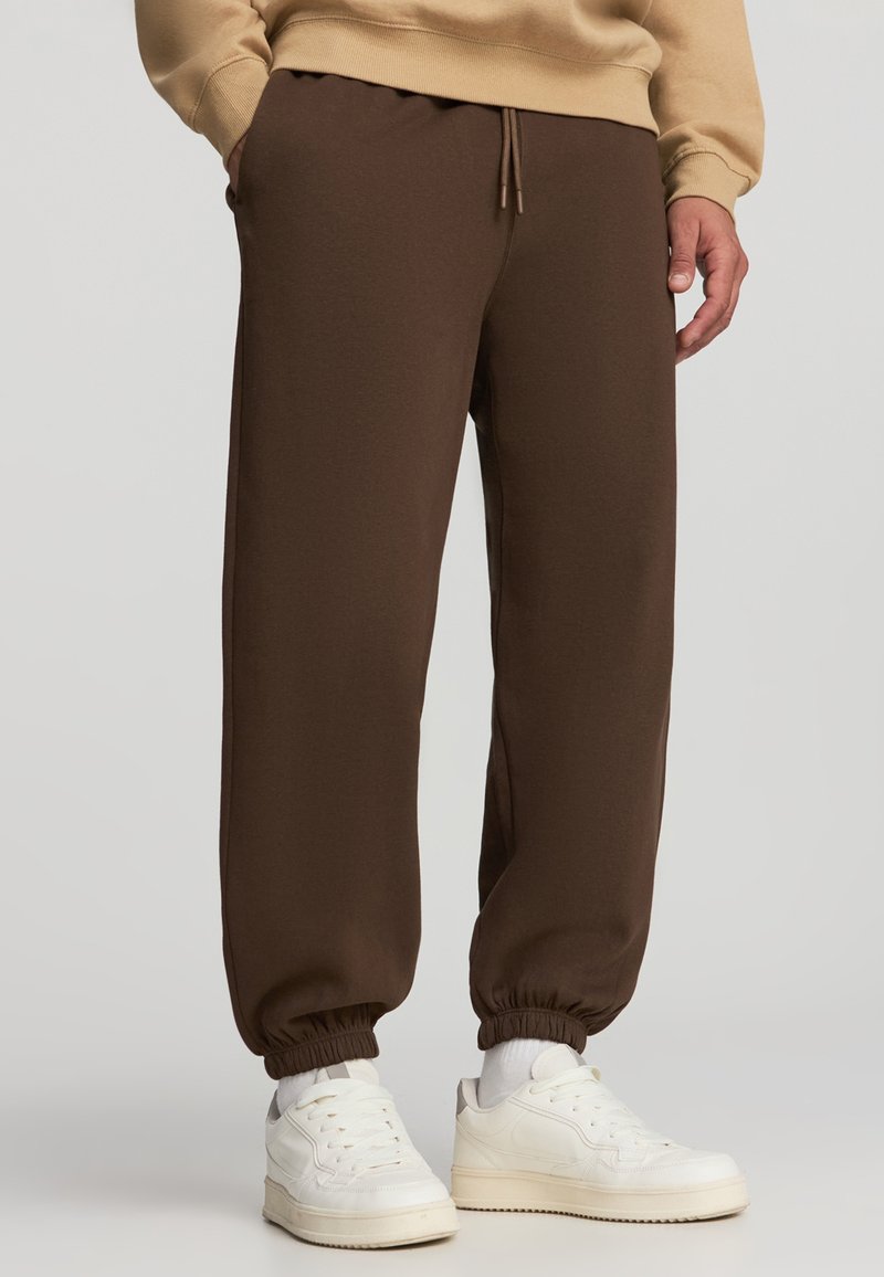 Brązowe dresy z elastycznym paskiem i ściągaczami, wykonane z miękkiego materiału, zestawione z białymi trampkami i beżową bluzą.