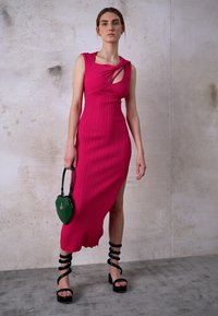 Vestido fúcsia com pregas, fenda lateral e detalhe recortado, combinado com uma bolsa verde em forma de coração e sandálias de salto alto pretas com tiras.