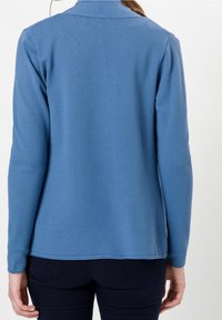 Top de manga larga azul con textura y cuello, que presenta un patrón de panal y un dobladillo recto, combinado con jeans oscuros.